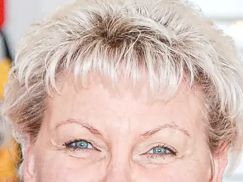 Eine lächelnde Frau mit kurzen blonden Locken und rotem Oberteil blickt in die Kamera.