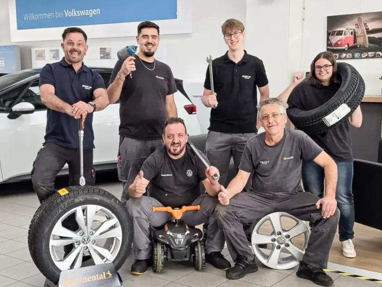 Sechs Werkstattmitarbeitende in schwarzen Poloshirts posieren mit Werkzeugen und Autoreifen in einem Autohaus.