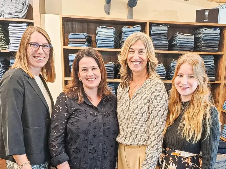 Vier lächelnde Frauen stehen nebeneinander in einem Bekleidungsgeschäft vor Regalen mit gefalteten Jeans.