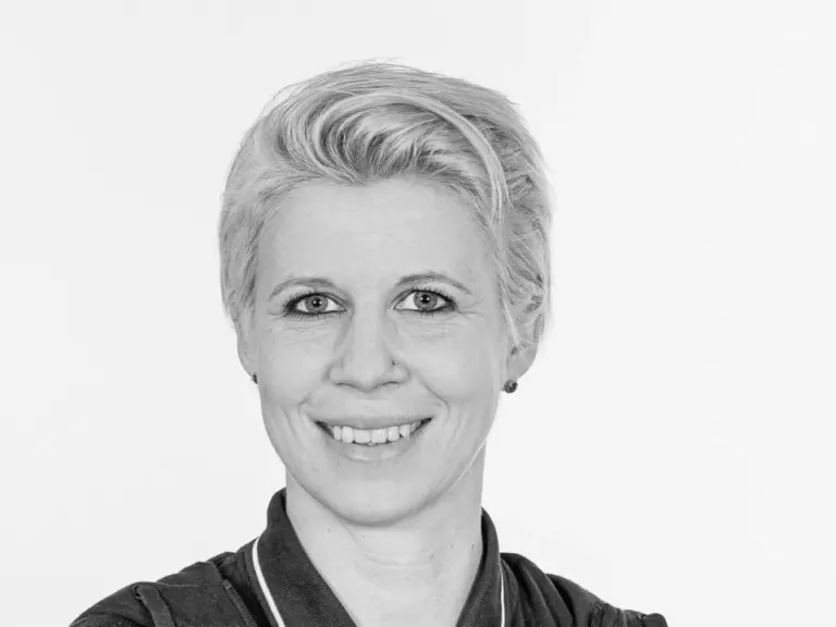 Frau mit kurzen grauen Haaren lächelt in Sportjacke vor hellem Hintergrund.