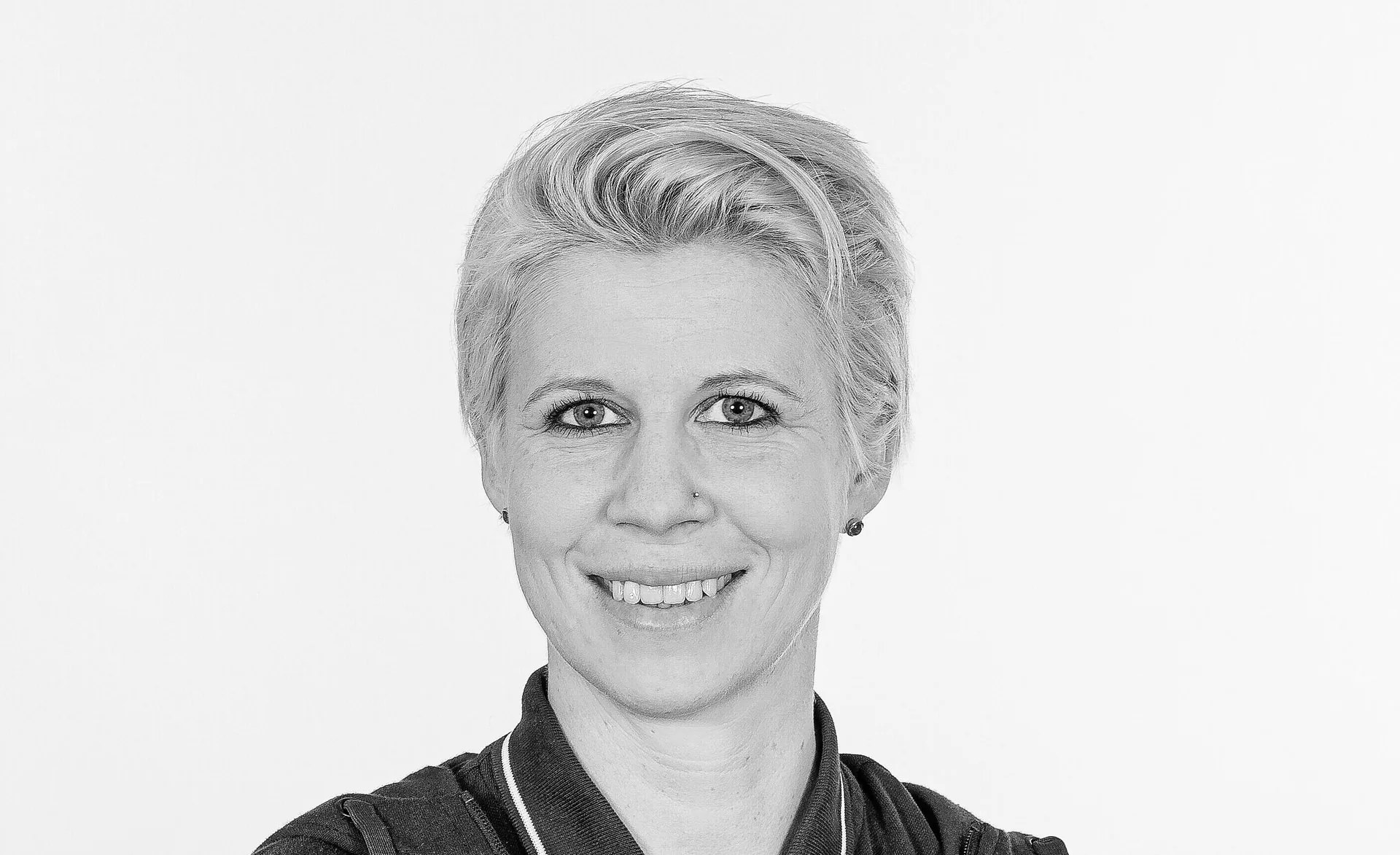 Frau mit kurzen grauen Haaren lächelt in Sportjacke vor hellem Hintergrund.