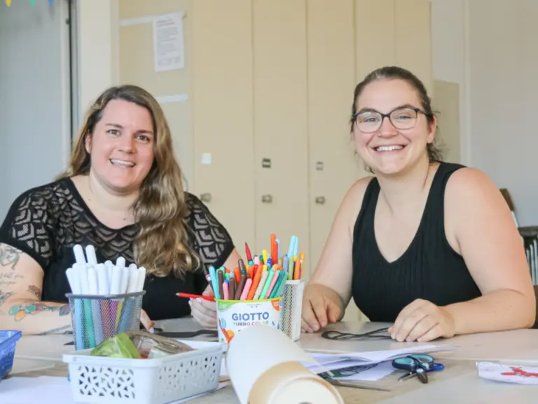 Zwei lächelnde Frauen sitzen an einem Tisch mit bunten Stiften, einer Papierrolle und Bastelmaterial.