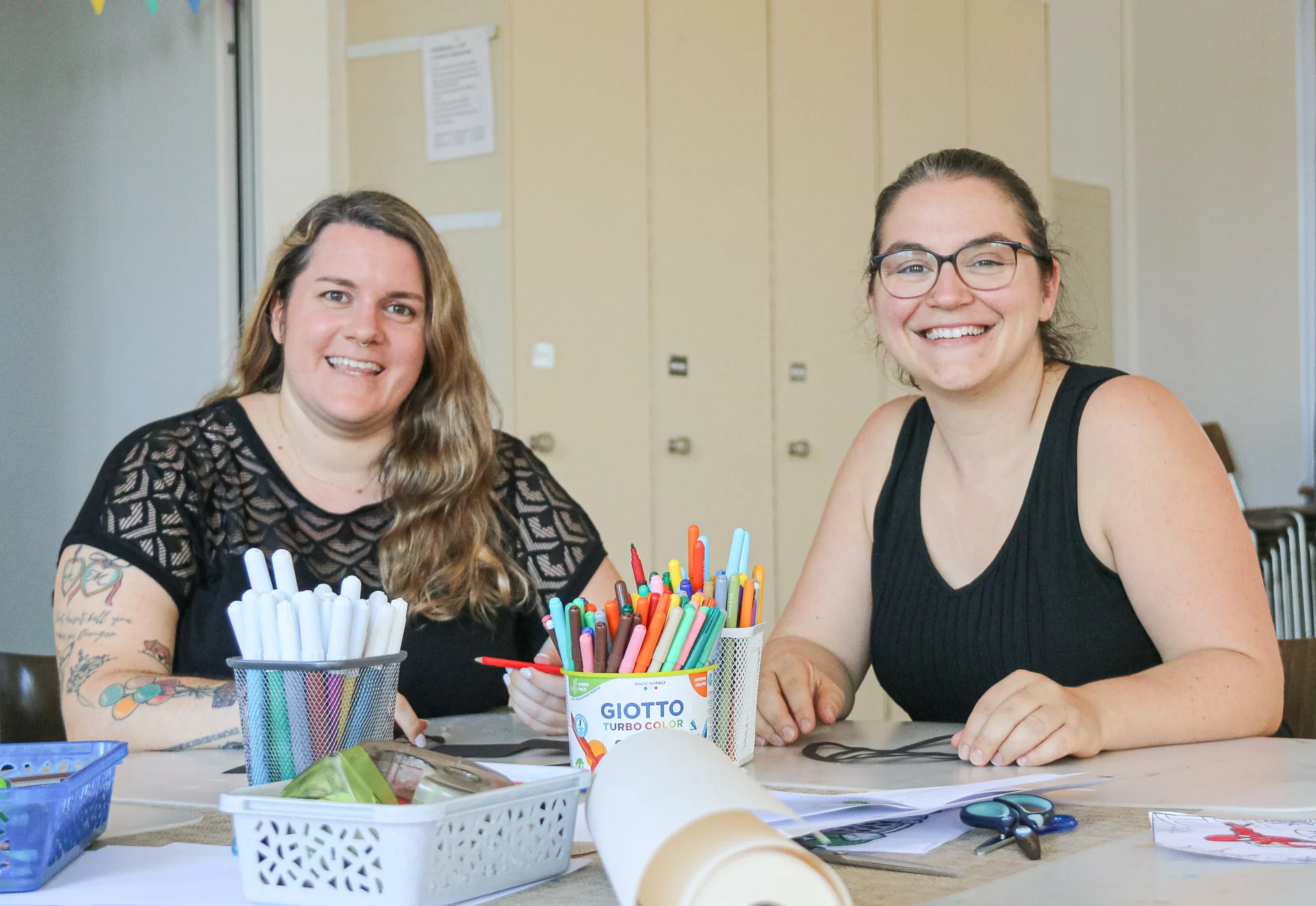 Zwei lächelnde Frauen sitzen an einem Tisch mit bunten Stiften, einer Papierrolle und Bastelmaterial.