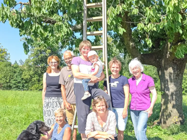 Gruppe von Frauen und ein Baby posiert vor einer Leiter unter einem Baum, ein Hund liegt daneben.