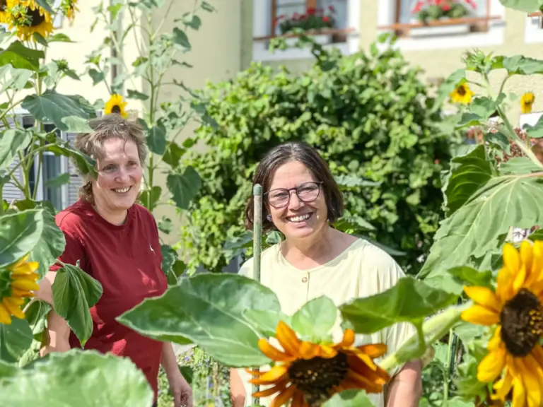 Zwei lachende Frauen stehen zwischen hohen Sonnenblumen in einem Garten vor einem Haus.