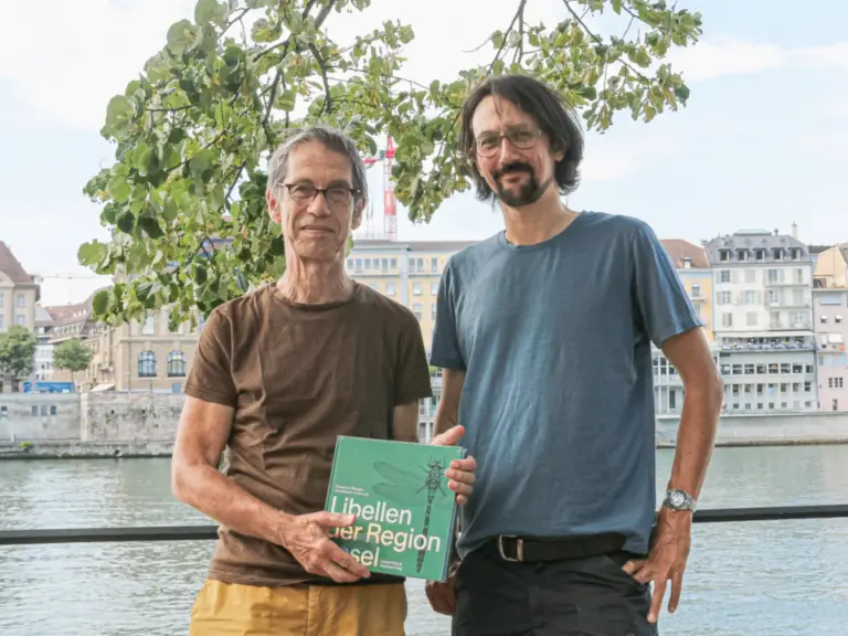 Zwei Männer stehen an einem Fluss vor einer Stadtkulisse, einer hält ein grünes Buch.