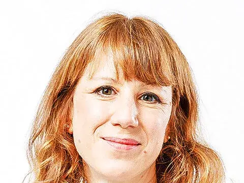 Frau mit schulterlangen roten Locken und Pony lächelt in die Kamera, dunkles Blazer, weisses Top.