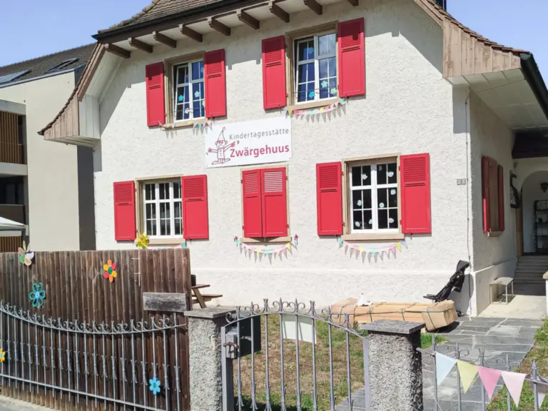 Weiss verputztes Haus mit rotem Ziegeldach, roten Fensterläden und bunten Wimpelketten hinter einem Zaun.