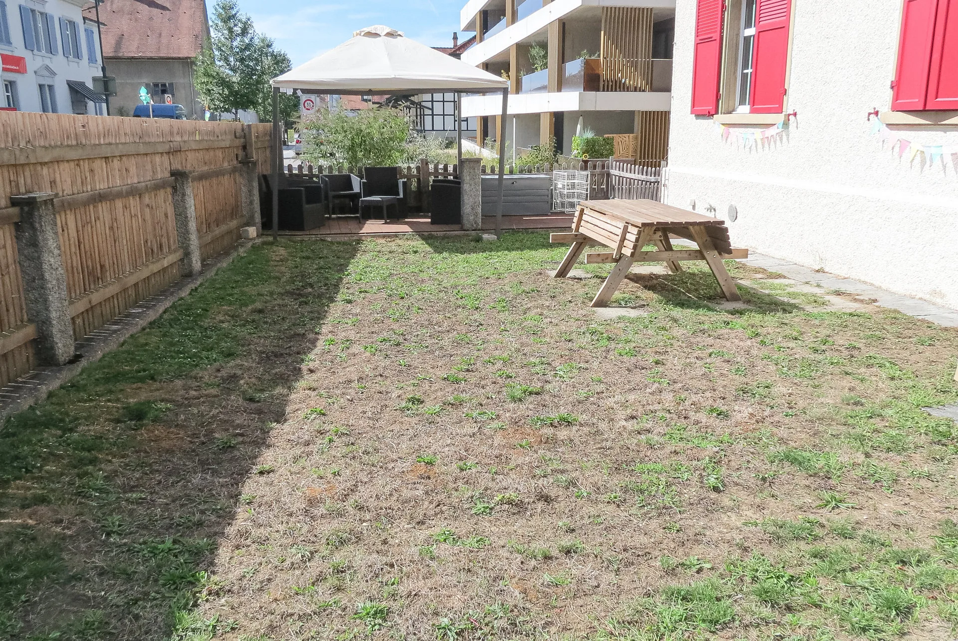 Ein Garten mit vertrocknetem Rasen, Holzzaun, Sitzgruppe, Pavillon und Picknicktisch neben einem Haus mit roten Fensterläden.