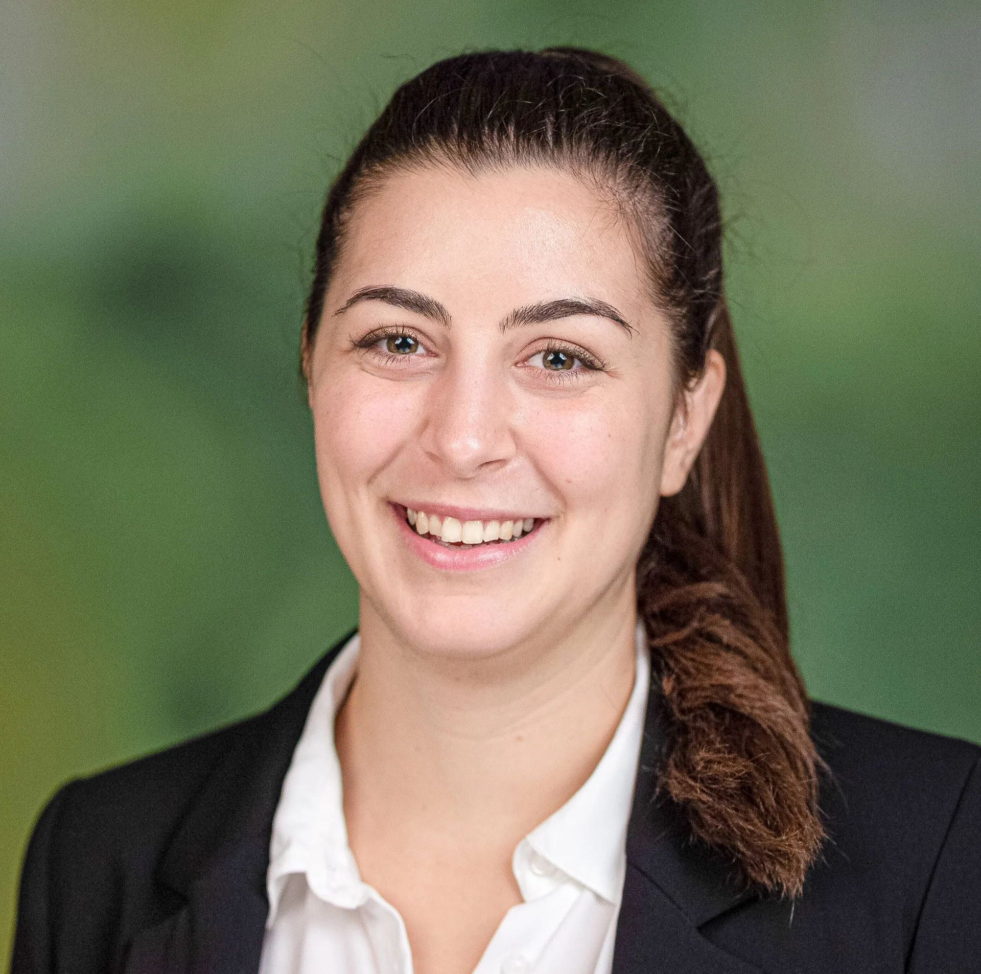 Lächelnde junge Frau mit zurückgebundenem braunem Haar, dunklem Blazer und weisser Bluse vor grünem Hintergrund.