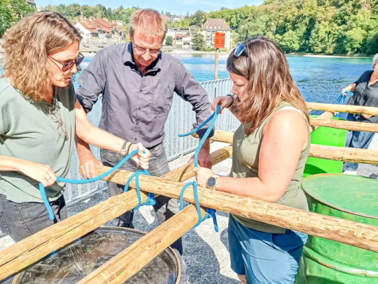 Drei Personen binden mit blauen Seilen Holzbalken an Fässern zu einem Floss zusammen.