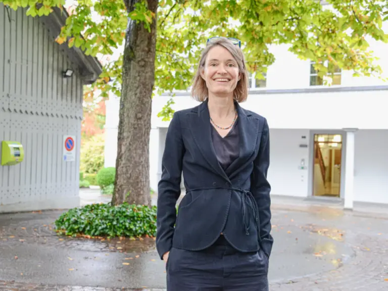 Eine lächelnde Frau mittleren Alters im dunklen Blazer steht auf einem Platz vor einem Baum.