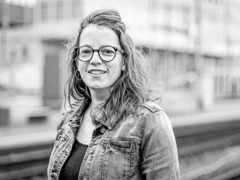 Lächelnde Frau mit Locken und Brille in Jeansjacke steht auf einem Bahnsteig, Schwarzweißfoto.