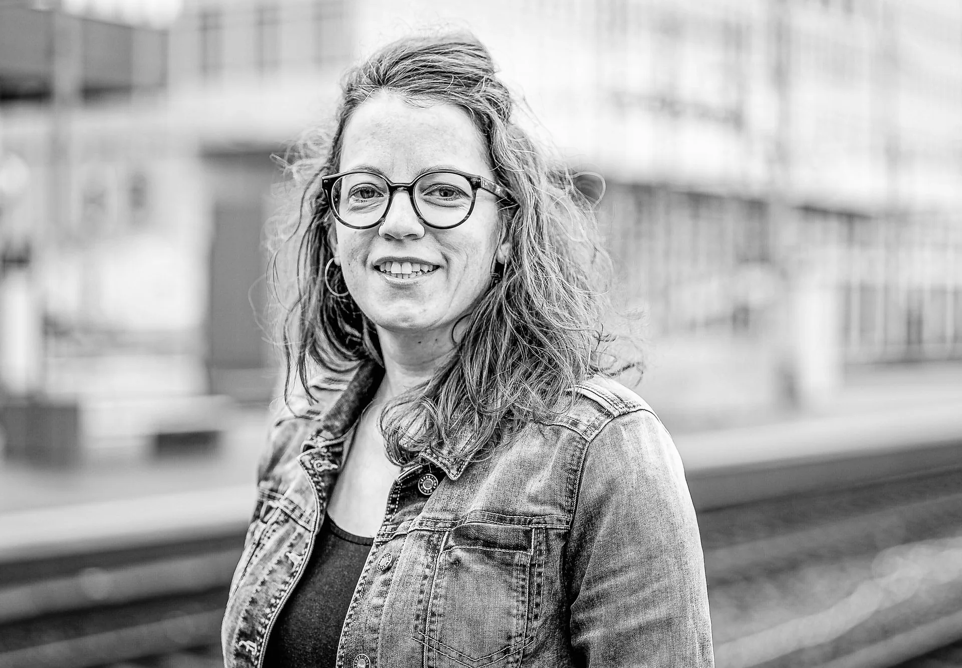 Lächelnde Frau mit Locken und Brille in Jeansjacke steht auf einem Bahnsteig, Schwarzweißfoto.