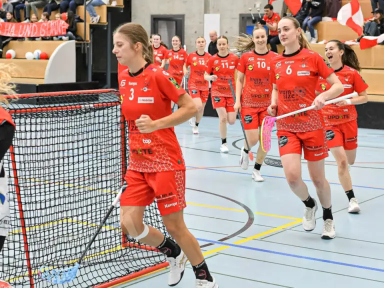 Unihockeyspielerinnen in roten Trikots laufen jubelnd in die Halle, im Hintergrund Fans mit Schweizer Fahnen.