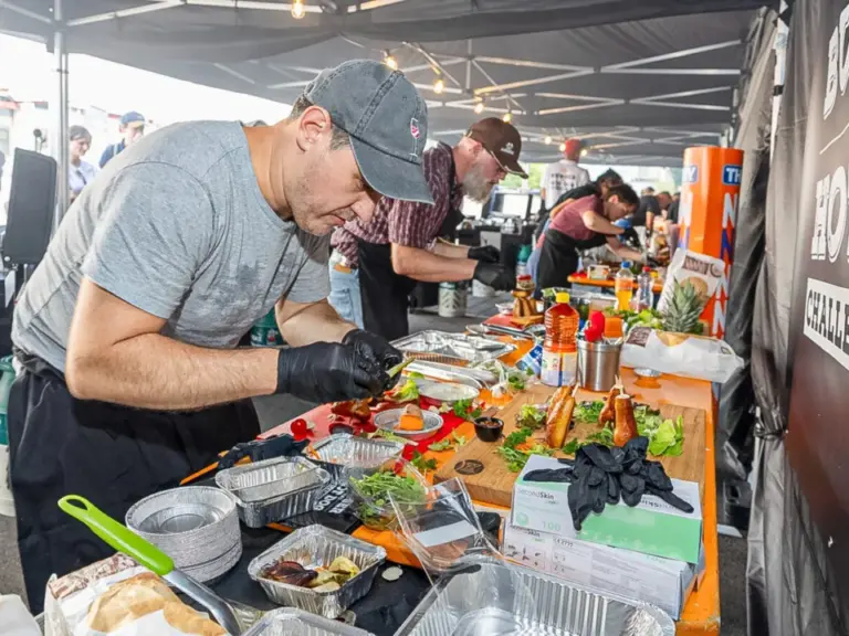 Männer mit Kappen bereiten unter einem Zelt an einem Stand Speisen zu, im Hintergrund ein Schild mit Burger-Wettbewerb.