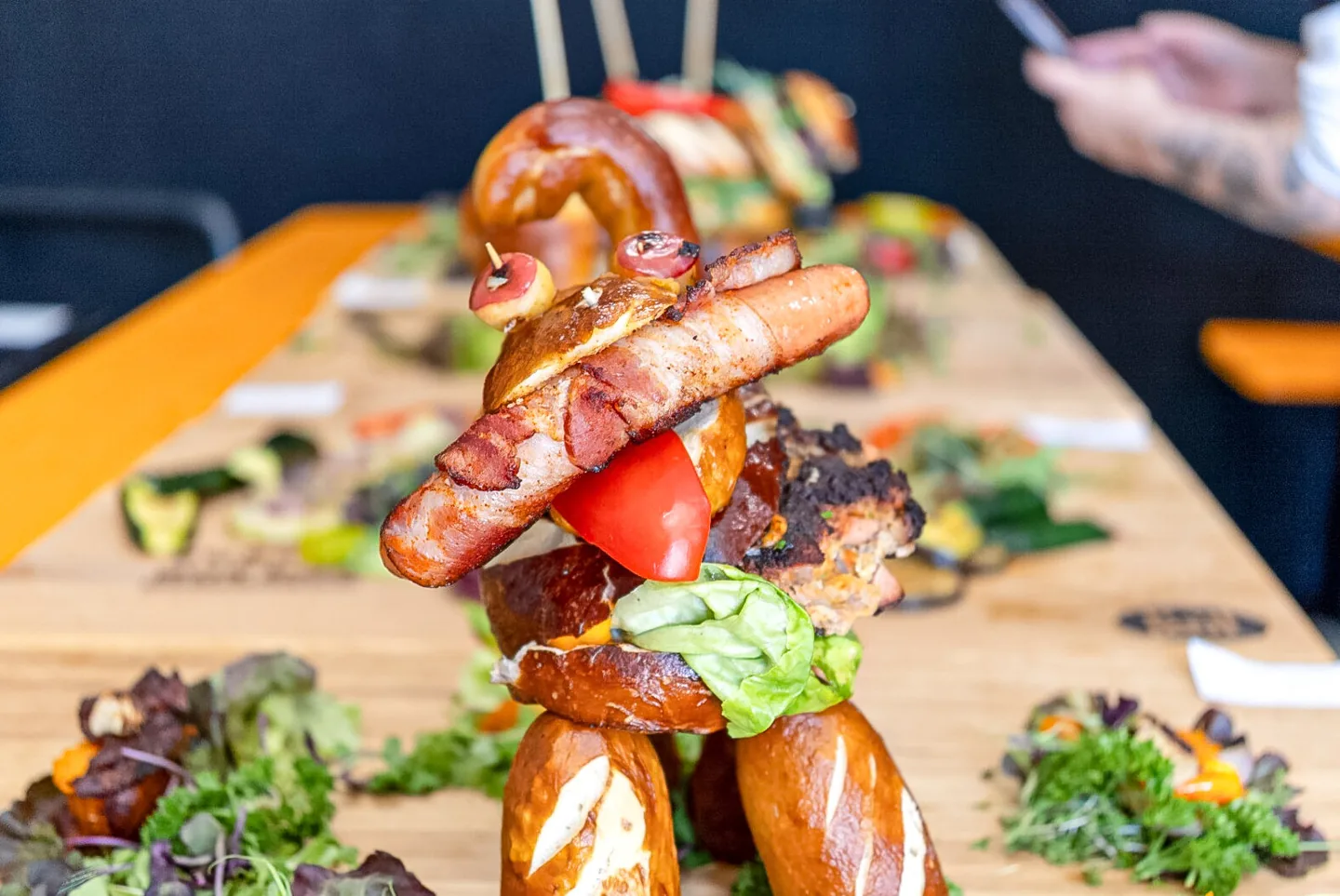 Ein aufwendig gestapelter Hotdog mit Bacon, Bratwurst, Salat und Brezel auf einem Holzbrett.