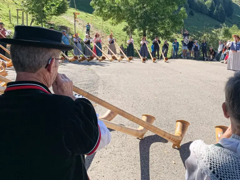 Alphornbläser in Tracht stehen im Halbkreis auf einer Strasse vor grünen Hügeln, Zuschauer im Hintergrund.