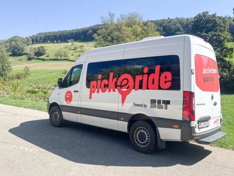 Ein weisser Kleinbus mit rotem «pick-e-ride»-Schriftzug steht auf einer Landstrasse vor grüner Hügellandschaft.