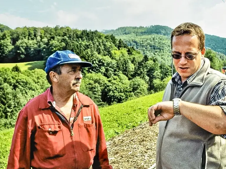 Zwei Männer stehen im Grünen vor bewaldeten Hügeln, einer schaut auf seine Armbanduhr, im Hintergrund eine Sesselliftstütze.