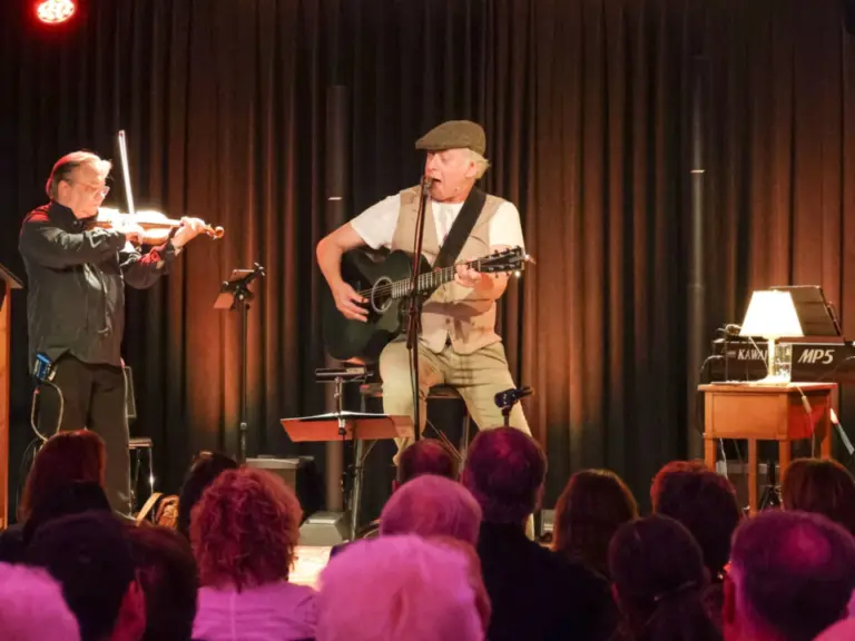 Drei Musiker auf einer Bühne spielen Geige, Gitarre und Keyboard vor sitzendem Publikum.