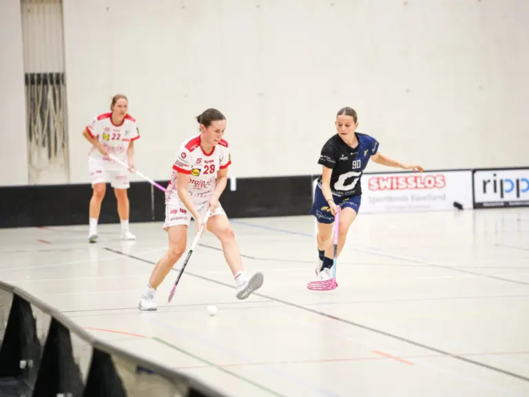 Drei Unihockeyspielerinnen laufen in einer Halle dem Ball hinterher, im Hintergrund Werbebanden.
