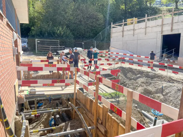 Eine Gruppe Menschen steht an einer Baugrube mit Rohrleitungen, umgeben von rot-weissen Absperrungen.