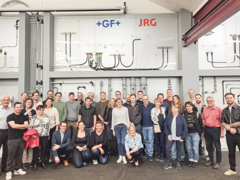 Grosse Gruppe von Menschen posiert für ein Gruppenfoto in einer Werkshalle mit Rohrleitungen.
