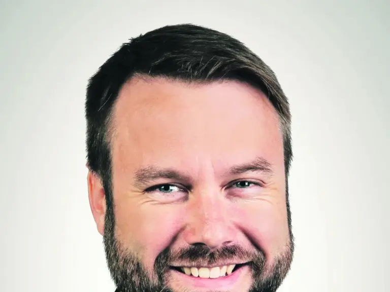 Lächelnder Mann mit Vollbart in Anzug und gestreifter Krawatte vor hellem Hintergrund.