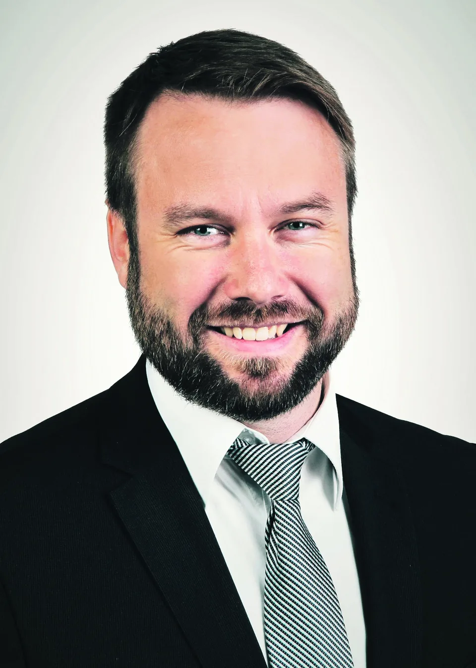 Lächelnder Mann mit Vollbart in Anzug und gestreifter Krawatte vor hellem Hintergrund.