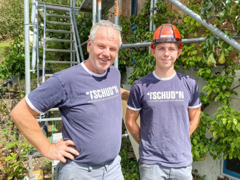 Zwei Männer in gleichen T-Shirts stehen lächelnd vor einem Baugerüst, einer trägt einen orangen Schutzhelm.