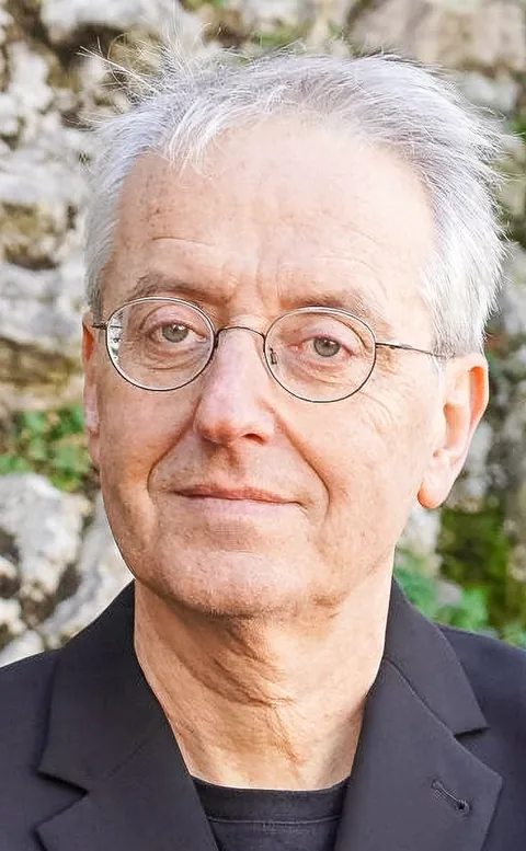 Ein älterer Mann mit grauen Haaren und runder Brille blickt ernst in die Kamera, im Hintergrund eine Steinmauer.