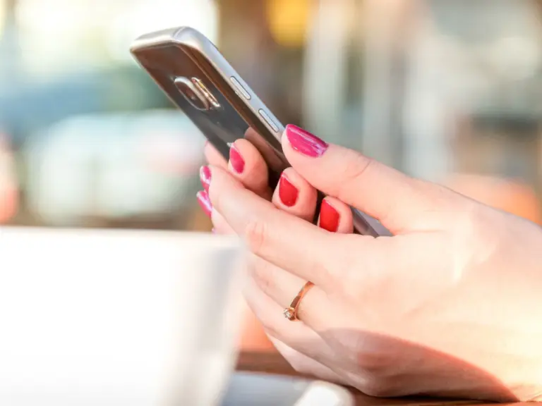 Eine Hand mit rot lackierten Nägeln hält ein Smartphone über einem Kaffeetisch im Freien.