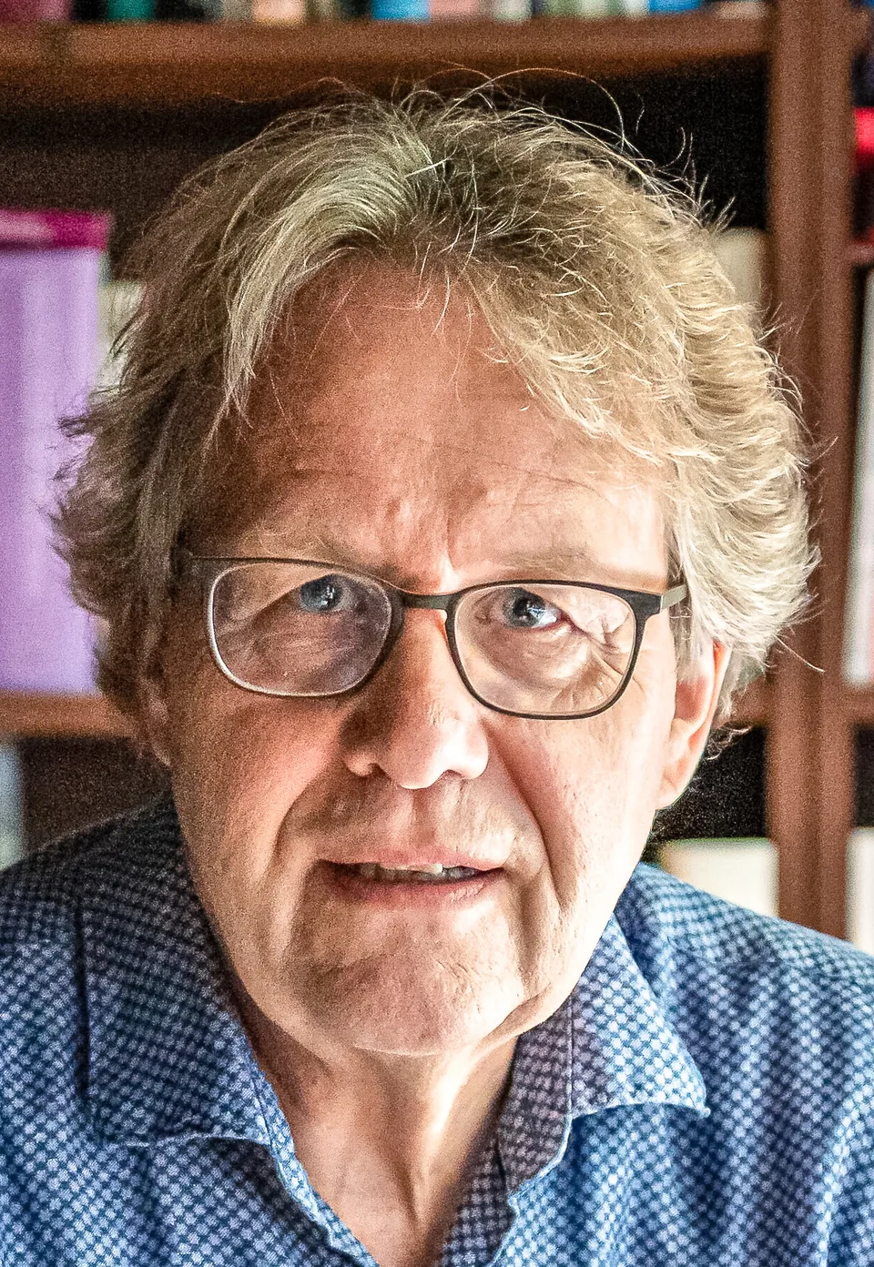 Ein älterer Mann mit Brille und grauem Haar blickt vor einem Bücherregal in die Kamera.