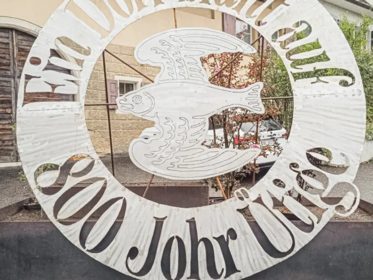 Ein rundes Metallschild mit Fischmotiv und der Aufschrift „Ein Dorf blüht auf, 800 Jahre“ vor einem Haus.