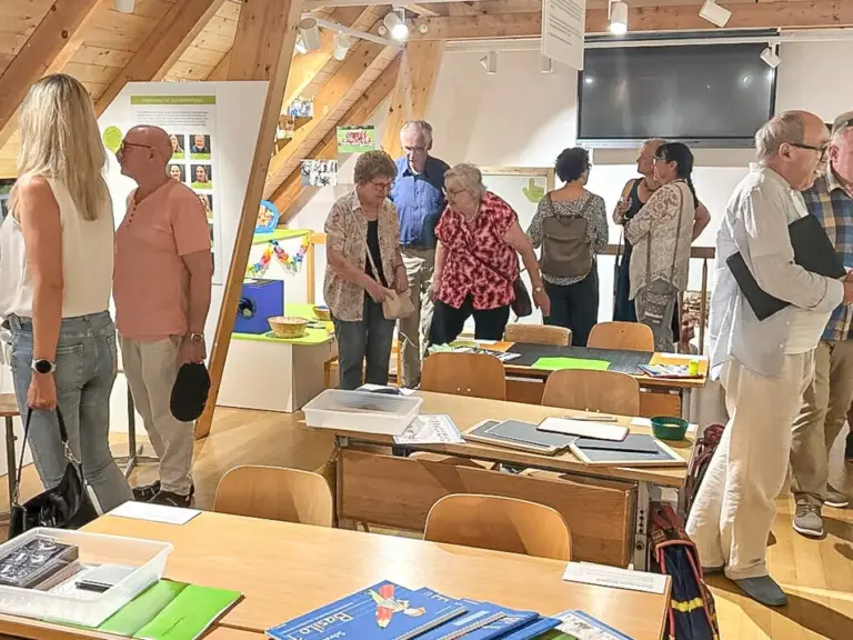 Eine Gruppe Erwachsener steht und unterhält sich in einem holzverkleideten Ausstellungsraum mit Tischen und Infotafeln.