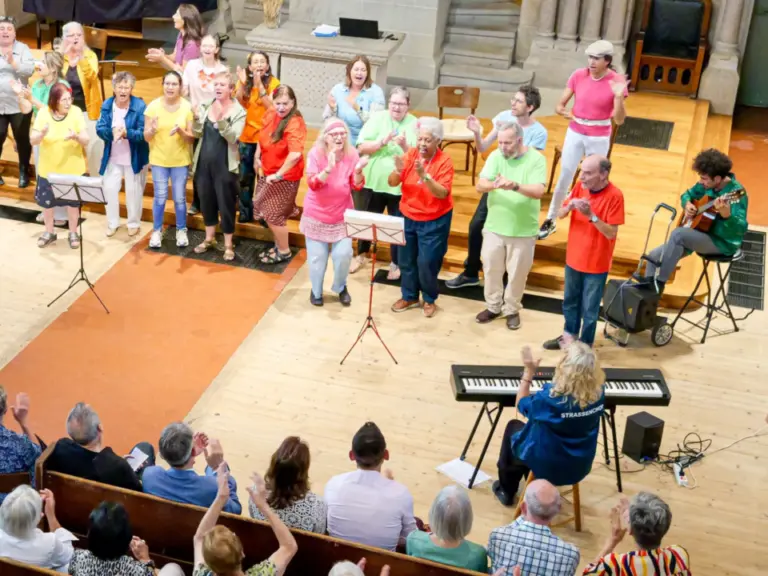 Chor in bunten Oberteilen singt und klatscht auf Kirchenstufen, davor Publikum in Bankreihen applaudiert.