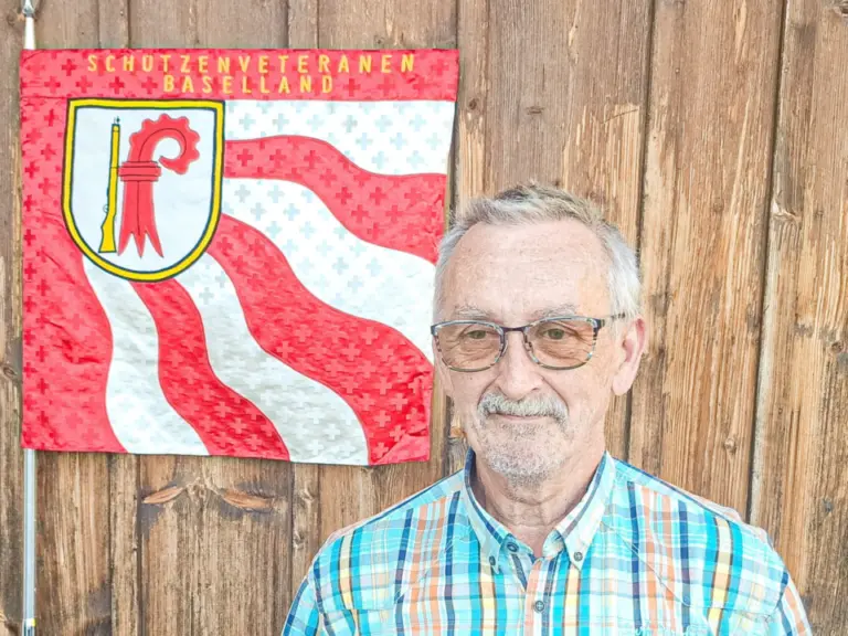 Älterer Mann mit Brille und kariertem Hemd steht vor einer Holzwand mit Wappenfahne.