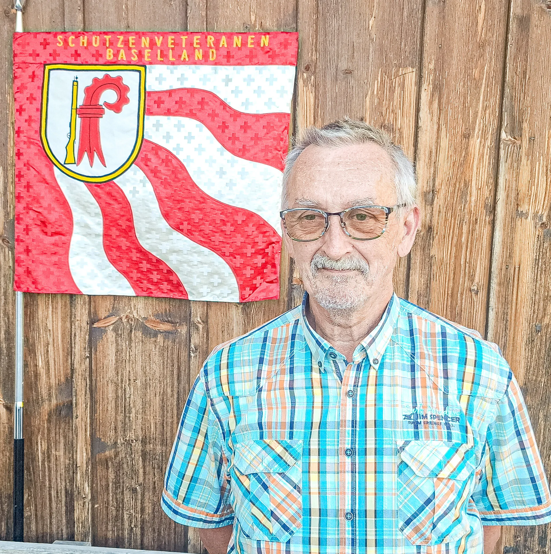 Älterer Mann mit Brille und kariertem Hemd steht vor einer Holzwand mit Wappenfahne.