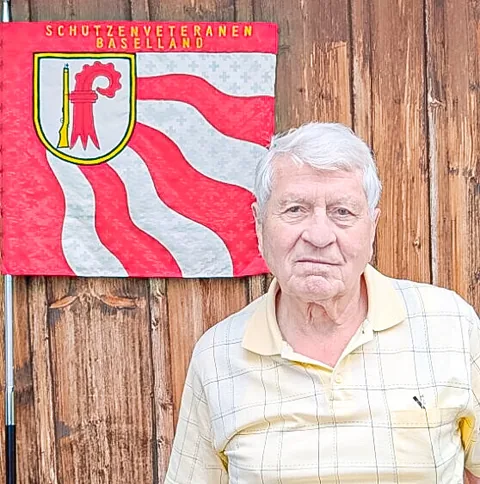 Ein älterer Mann mit weissem Haar in kariertem Hemd steht vor einer Holzwand neben einer Vereinsfahne.