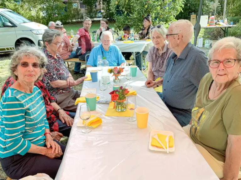Eine Gruppe älterer Menschen sitzt an einem langen, weiss gedeckten Tisch im Freien und unterhält sich.