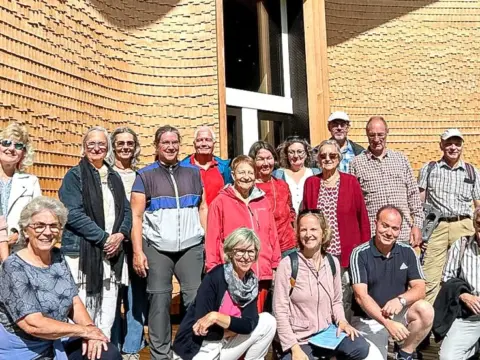 Eine Gruppe von Menschen posiert lächelnd vor einer geschwungenen Holzfassade eines modernen Gebäudes.