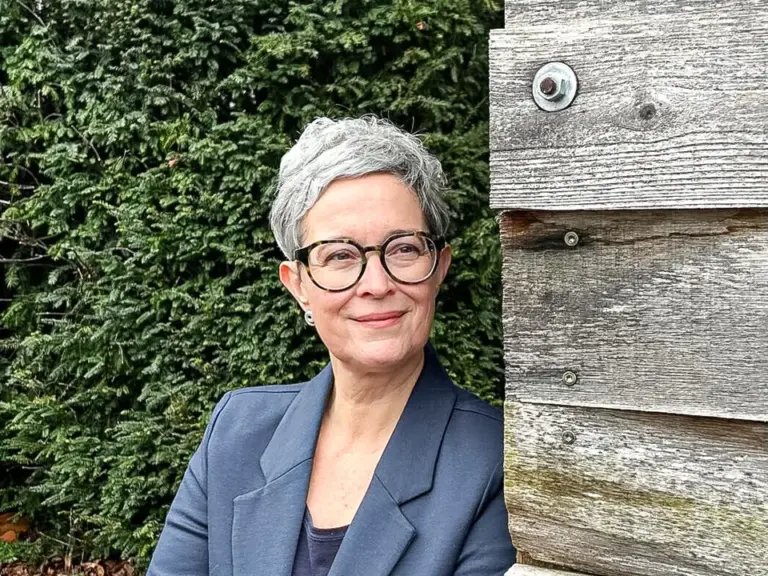 Eine Frau mittleren Alters mit kurzen grauen Haaren und Brille lehnt lächelnd an einer Holzwand im Garten.