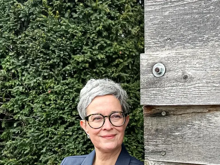 Eine Frau mit grauen kurzen Haaren und Brille lehnt lächelnd an einer Holzwand, Arme verschränkt.