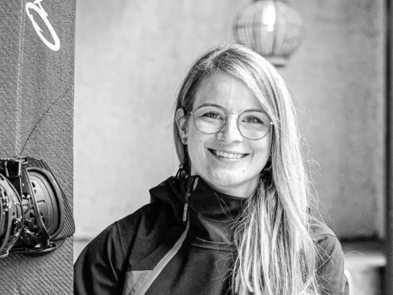 Lächelnde langhaarige Frau in Jacke sitzt neben einem Snowboard auf einer Holzbank, Schwarzweissfoto.