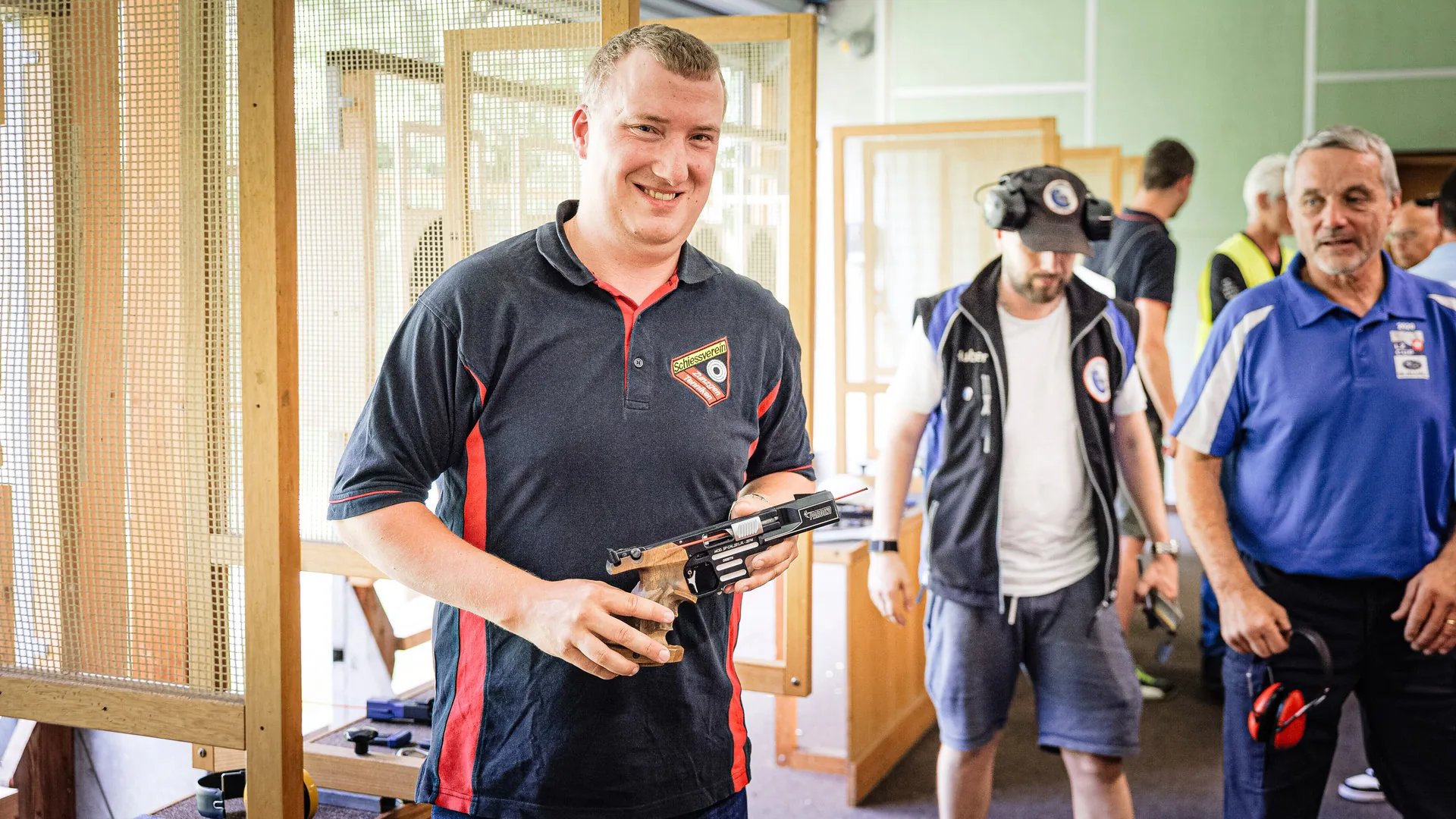 Ein lächelnder Mann hält eine Sportpistole in einer Schiesshalle, im Hintergrund weitere Personen.