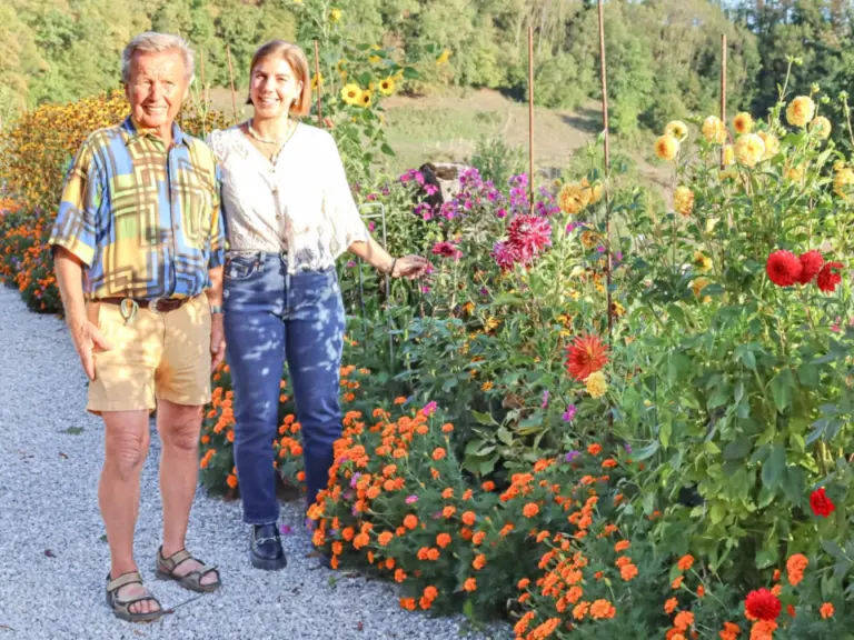 Ein älterer Mann und eine jüngere Frau stehen lächelnd nebeneinander an einem Weg entlang einer bunten Blumenrabatte.