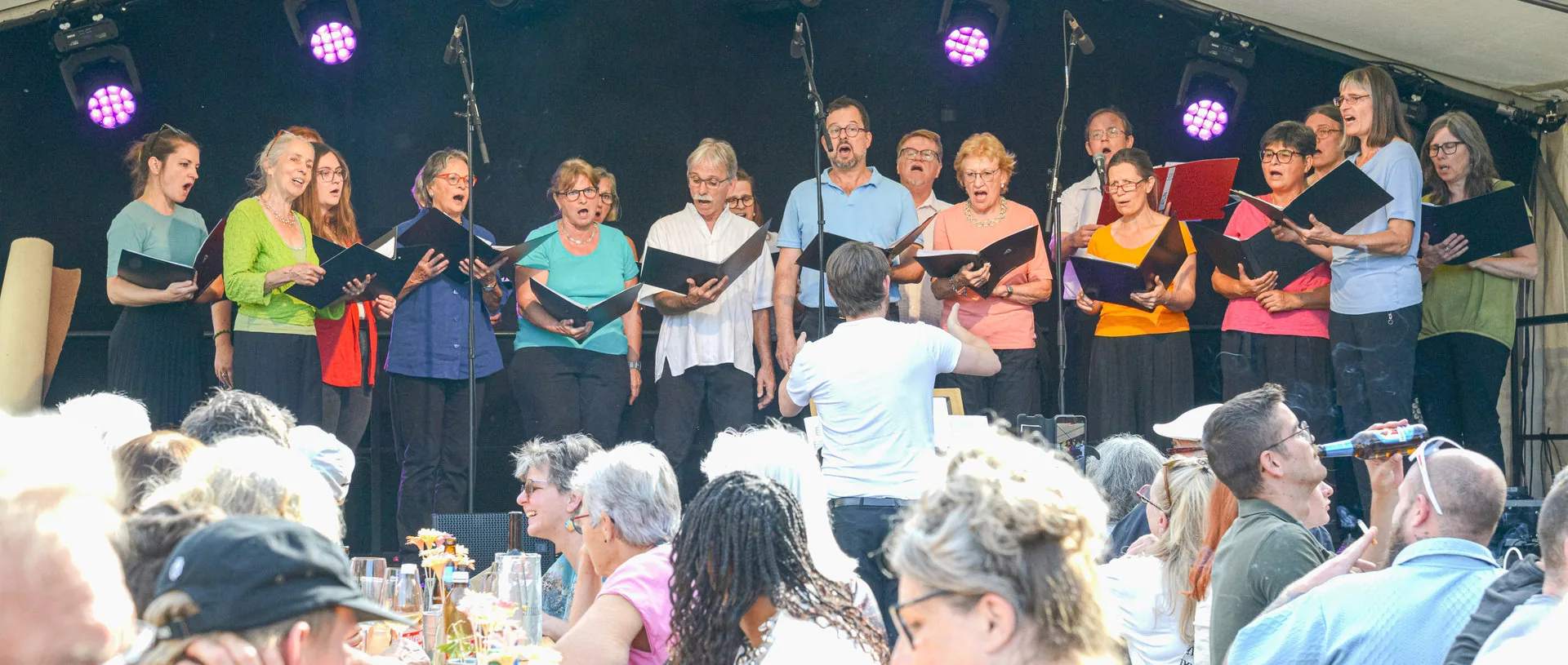 Chormitglieder mit Notenmappen singen auf einer Bühne, davor sitzt Publikum an Tischen.