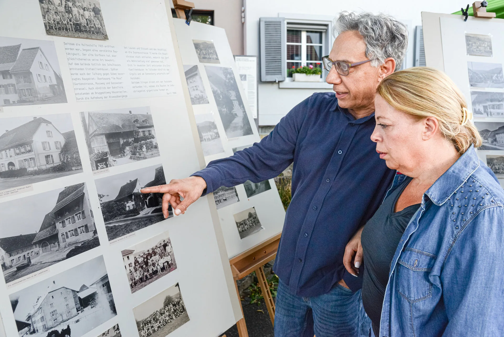 Ein Mann und eine Frau betrachten alte Schwarzweissfotos von Häusern auf ausgestellten Tafeln.
