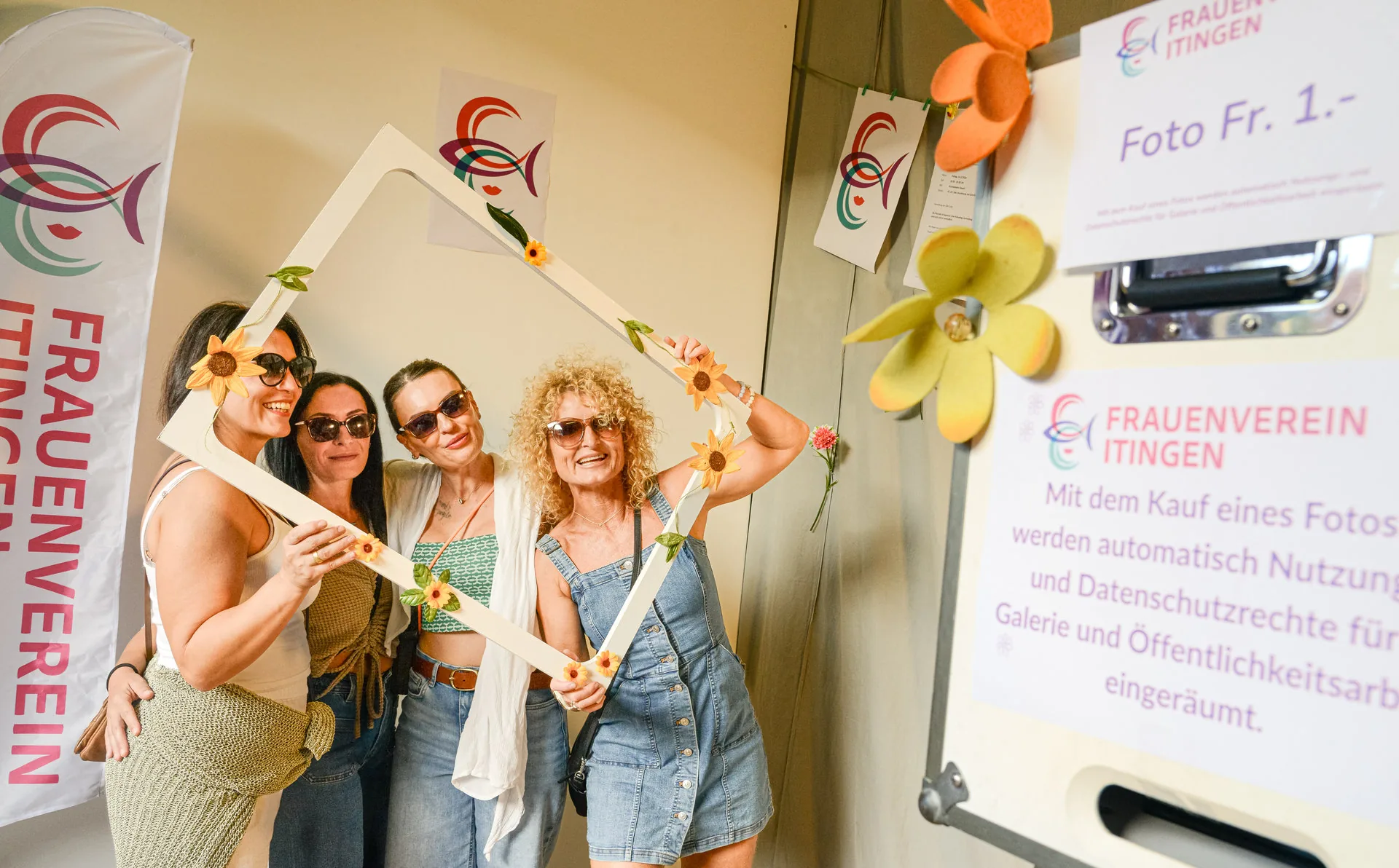 Vier Frauen mit Sonnenbrillen posieren lachend hinter einem dreieckigen Fotorahmen mit Sonnenblumen.
